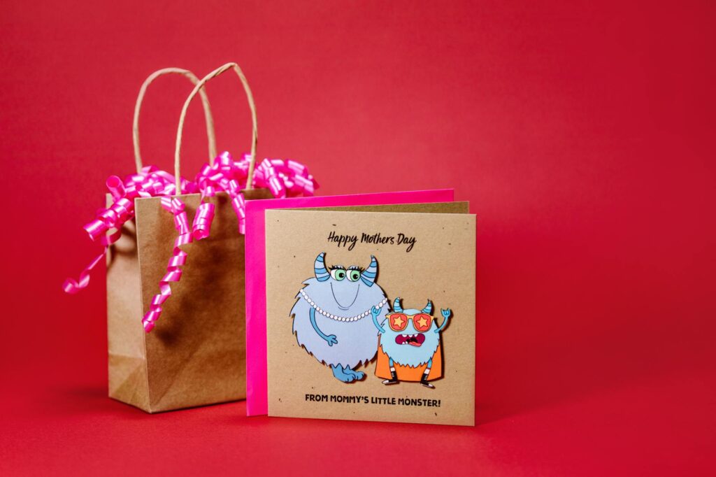 Charming Mother's Day card featuring cute monsters and a gift bag on a red background.